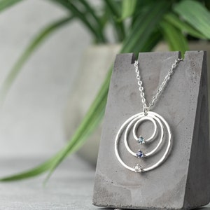 May include: A silver necklace with three concentric circles and three small gemstones. The necklace is on a gray display stand.