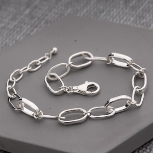 May include: A silver-toned chain bracelet featuring elongated, oval-shaped links with a textured, hammered finish. The bracelet includes a secure lobster claw clasp and a short extender chain ending in a small, smooth spherical bead detail.