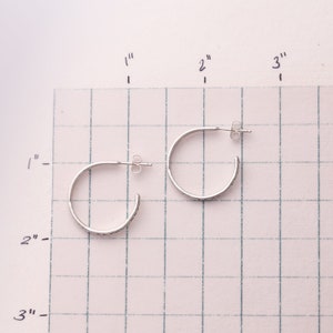 Pode incluir: Dois brincos de argola de prata com um design texturizado. Os brincos s&atilde;o mostrados em uma superf&iacute;cie quadriculada com medidas em polegadas.