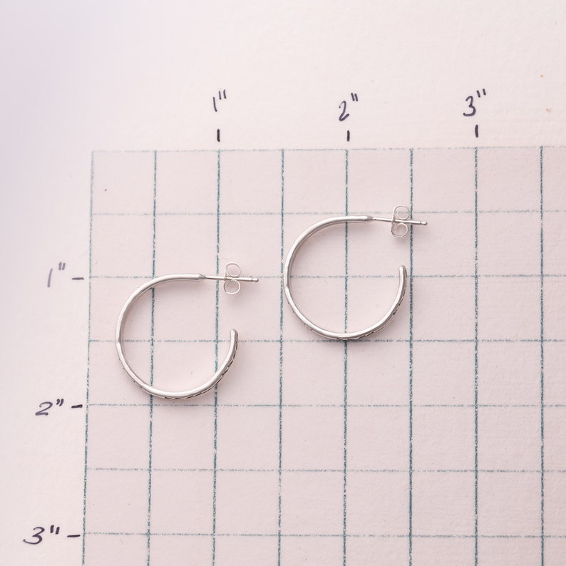 Pode incluir: Dois brincos de argola de prata com um design texturizado. Os brincos s&atilde;o mostrados em uma superf&iacute;cie quadriculada com medidas em polegadas.
