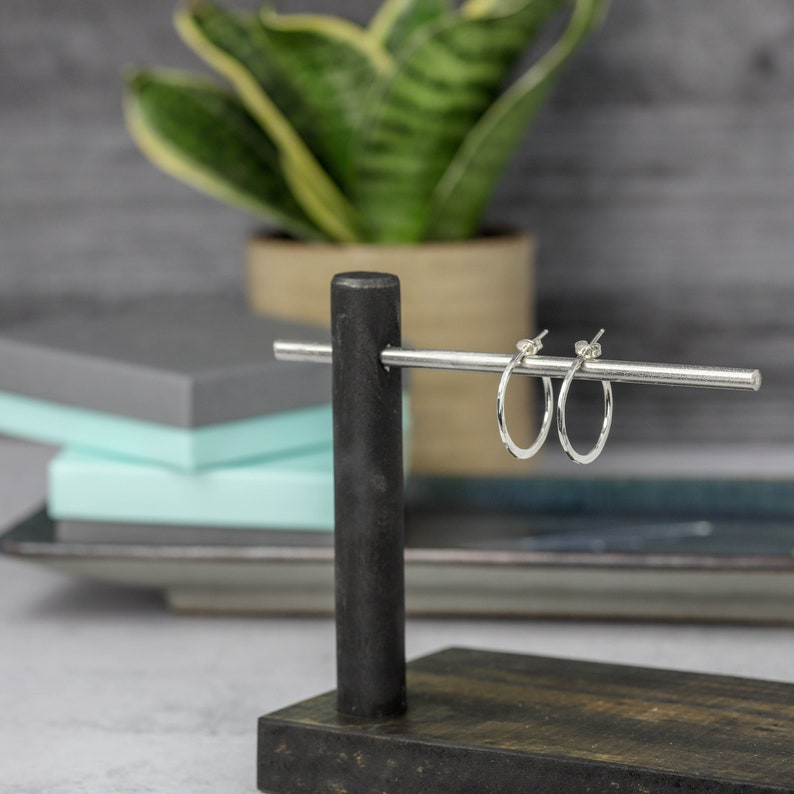 A pair of sterling silver hoops hang from a wooden jewelry display.  This angle shows that they are narrow from the front and all sides have a rustic hammered texture.  The earrings are polished to a high reflective shine and measure one inch long