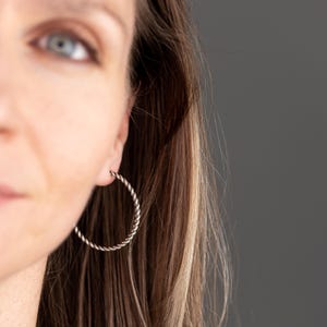 May include: Silver-toned hoop earrings with a twisted rope design. The earrings are circular and worn on the ear. The image shows a close-up of the earring and part of the person's face and hair.