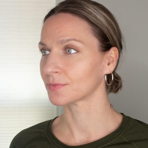 K&ouml;nnte beinhalten: Eine Frau mit hellbraunen Haaren, die ein dunkelgr&uuml;nes Shirt und silberne Creolen tr&auml;gt.