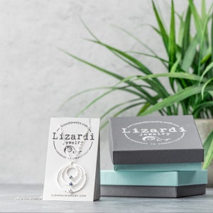 May include: A silver necklace with a circle design and two blue gemstones. The necklace is displayed on a white card with the text "Lizardi Jewelry" and "Handmade in Connecticut". The necklace is also displayed in a gray box with the text "Lizardi Jewelry" and "Handmade in Connecticut".
