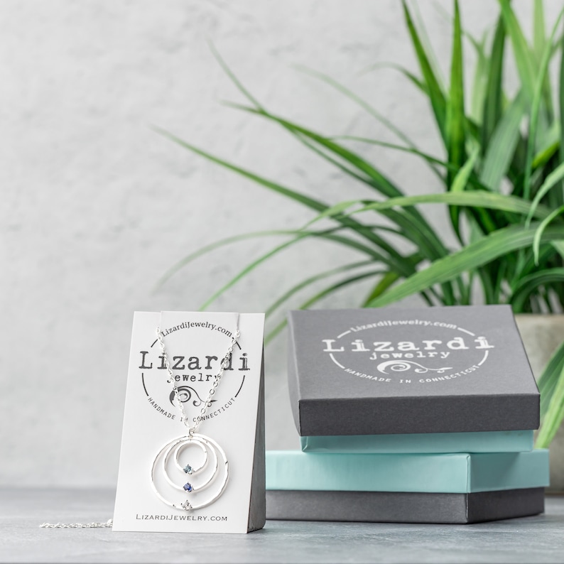 May include: A silver necklace with a circle design and two blue gemstones. The necklace is displayed on a white card with the text "Lizardi Jewelry" and "Handmade in Connecticut". The necklace is also displayed in a gray box with the text "Lizardi Jewelry" and "Handmade in Connecticut".