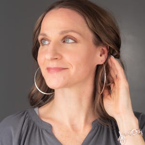 May include: Large, thin, circular silver hoop earrings with a polished, reflective finish. The earrings are simple, circular bands of metal that hang from the earlobes, creating a wide, open loop design that frames the face.