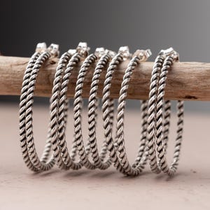 May include: A collection of silver-colored hoop earrings with a twisted rope design. The earrings are displayed on a wooden dowel against a blurred background. The earrings are a medium size.