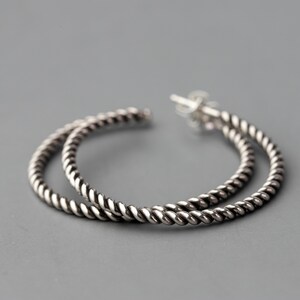 May include: A pair of silver-colored hoop earrings with a twisted rope design. The earrings are circular and have a post closure. The background is a solid gray.