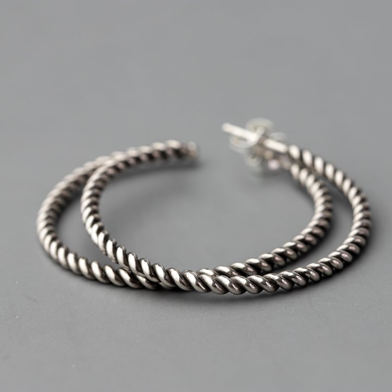May include: A pair of silver-colored hoop earrings with a twisted rope design. The earrings are circular and have a post closure. The background is a solid gray.