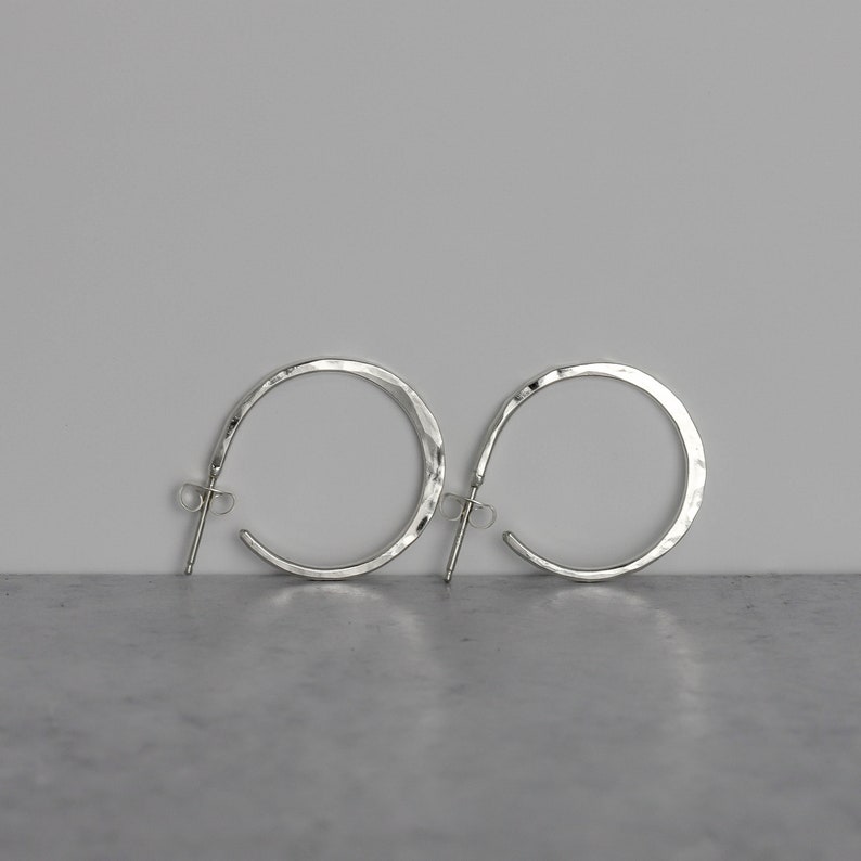 Hammered sterling silver hoops earrings side by side against a white background.  The side view of these once inch silver hoops shows the hand hammered texture and accentuates the fact that they are slightly flared at the center of the hoop.