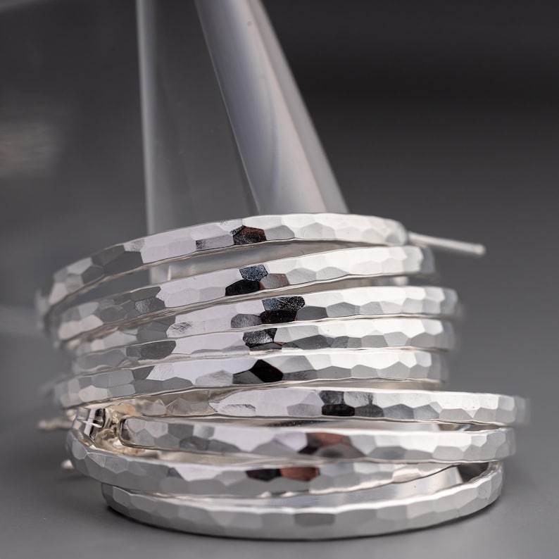 Close-up of several hammered sterling silver hoops stacked together. The hoops have a textured, faceted surface and are displayed on a stand.  Each hoop is thick and substantial, showcasing a unique hammered metal design. The silver has a bright, polished finish. The image highlights the handcrafted quality and the metallic sheen of the jewelry.