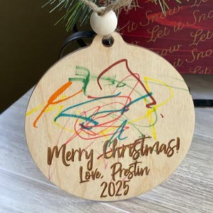 May include: A round wooden Christmas ornament with colorful scribbles and the words "Merry Christmas! Love, Prestin 2025" engraved. The ornament has a hole at the top with a twine hanger and a wooden bead.
