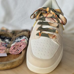 May include: Cream-coloured trainers with patterned ribbon laces. The shoe has a perforated toe box and a beige trim. A bundle of patterned fabric is visible on a wooden tray. The laces have a green, brown, and orange design.