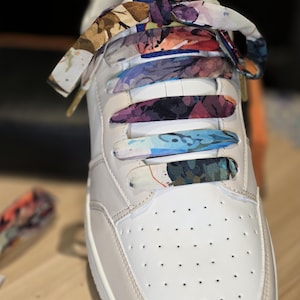 May include: White leather trainer with floral patterned laces. The laces feature a colourful design with shades of purple, blue, and orange. The shoe has a classic design with a perforated toe box.