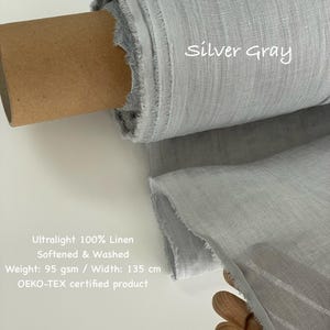 May include: A roll of silver-gray ultralight 100% linen fabric. The fabric is softened and washed, with a weight of 95 gsm and a width of 135 cm. The text "Silver Gray" is written on the fabric. The fabric is an OEKO-TEX certified product.