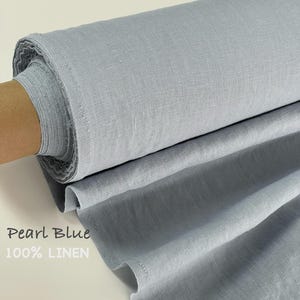 May include: A bolt of pearl blue fabric made of 100% linen. The material is wrapped around a brown cardboard core, showing a visible woven texture and a soft, cool-toned blue color. The text Pearl Blue 100% LINEN is printed on the fabric.