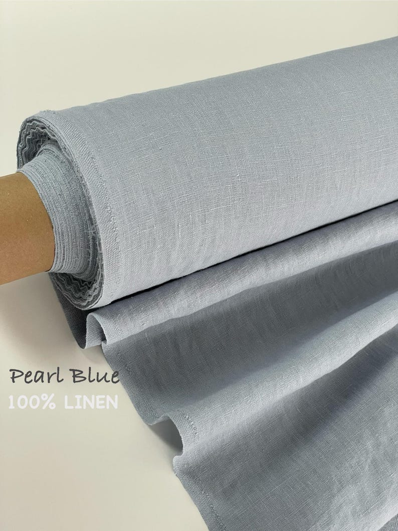 May include: A bolt of pearl blue fabric made of 100% linen. The material is wrapped around a brown cardboard core, showing a visible woven texture and a soft, cool-toned blue color. The text Pearl Blue 100% LINEN is printed on the fabric.