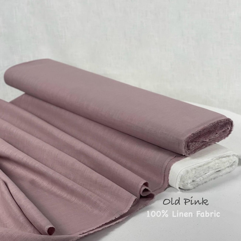 May include: A roll of old pink 100% linen fabric with a visible woven texture. The fabric is draped in soft folds, showcasing the dusty rose color and the natural weave of the textile material. The text Old Pink 100% Linen Fabric is visible.
