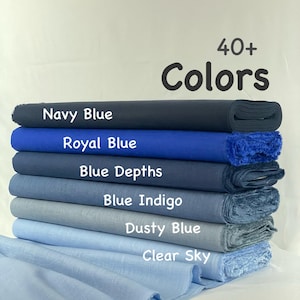 K&ouml;nnte beinhalten: Ein Stapel aus sechs gefalteten Stoffballen in verschiedenen Blaut&ouml;nen, beschriftet mit dem Text 40+ Colors, Navy Blue, Royal Blue, Blue Depths, Blue Indigo, Dusty Blue und Clear Sky, die einen Farbverlauf von Dunkel- zu Hellblau zeigen.