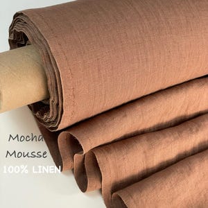Puede incluir: Un rollo de tela 100% lino de color mousse de moca. El tejido presenta una textura tejida visible y está drapeado en pliegues suaves. El texto "Mocha Mousse 100% LINEN" está impreso en el lado izquierdo del rollo de tela en fuente gris oscuro.