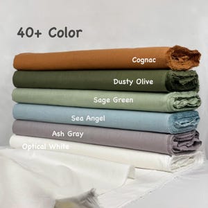 Puede incluir: Una pila de seis rollos de tela doblados en varios colores etiquetados como 40+ Color, Cognac, Dusty Olive, Sage Green, Sea Angel, Ash Gray y Optical White. Cada rollo presenta un tejido texturizado con bordes deshilachados, apilados horizontalmente.