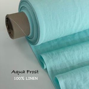 May include: A bolt of aqua frost colored fabric made of 100% linen. The textile is wrapped around a cardboard core, showing a visible woven texture and a light turquoise hue. The text Aqua Frost 100% LINEN is printed on the lower left side.