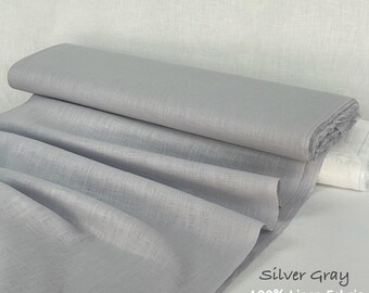 May include: A bolt of silver gray 100% linen fabric with a visible woven texture. The material is folded in layers, creating soft drapes and shadows across the surface. The text Silver Gray 100% Linen Fabric is printed in the bottom corner.