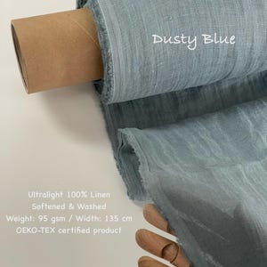 May include: A roll of dusty blue ultralight 100% linen fabric. The fabric is softened and washed, with a weight of 95 gsm and a width of 135 cm. The text "Dusty Blue" is written on the fabric. The product is OEKO-TEX certified.