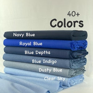 May include: A stack of six fabric bolts in various shades of blue, labeled Navy Blue, Royal Blue, Blue Depths, Blue Indigo, Dusty Blue, and Clear Sky. The text 40+ Colors is displayed above the fabric stack, which features a textured weave.