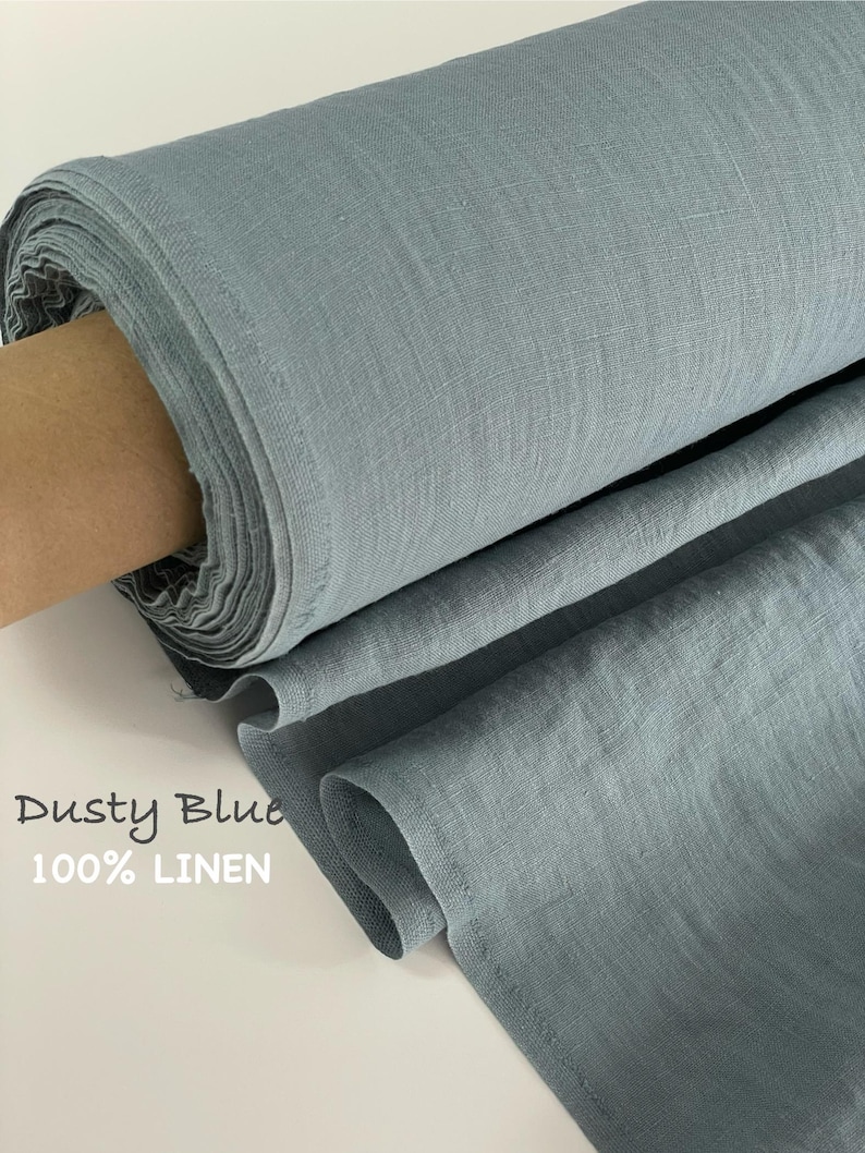 May include: A roll of dusty blue 100% linen fabric with a visible woven texture. The fabric is tightly wound on a cardboard core, with a portion of the material unrolled to show the drape and color. The text Dusty Blue 100% LINEN is visible.