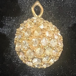May include: A spherical pendant featuring a gold-toned metal setting encrusted with numerous clear, round-cut faceted stones. The charm includes a small, triangular-shaped bail at the top for attachment, creating a textured, reflective surface.