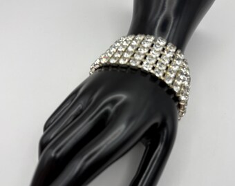 May include: A wide, silver-toned bracelet with multiple rows of clear, sparkling rhinestones. The bracelet is displayed on a black hand-shaped jewelry holder against a white background. The bracelet is a statement piece.