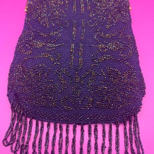 May include: A vintage beaded handbag in deep purple with a gold-toned metal frame. The bag features an intricate beaded pattern and a fringe along the bottom. The bag is approximately 8 inches in height.