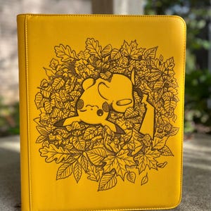 May include: A bright yellow binder featuring a detailed black line art illustration of a Pikachu character lying on its back, surrounded by a dense circular arrangement of various autumn leaves and small forest foliage.