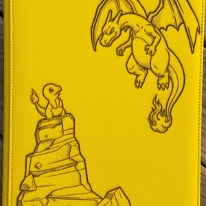 May include: A bright yellow rectangular binder featuring a dark brown laser-engraved illustration of a small lizard-like creature sitting on a rocky formation and a large winged dragon flying above it. The word Vaultx is etched at the bottom.