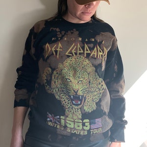 Puede incluir: Sudadera negra con efecto blanqueado que presenta un gr&aacute;fico de un leopardo con el texto "Pyromania Def Leppard 1983 United States Tour" impreso en la parte delantera. La sudadera la lleva puesta una persona que lleva una gorra de b&eacute;isbol marr&oacute;n.