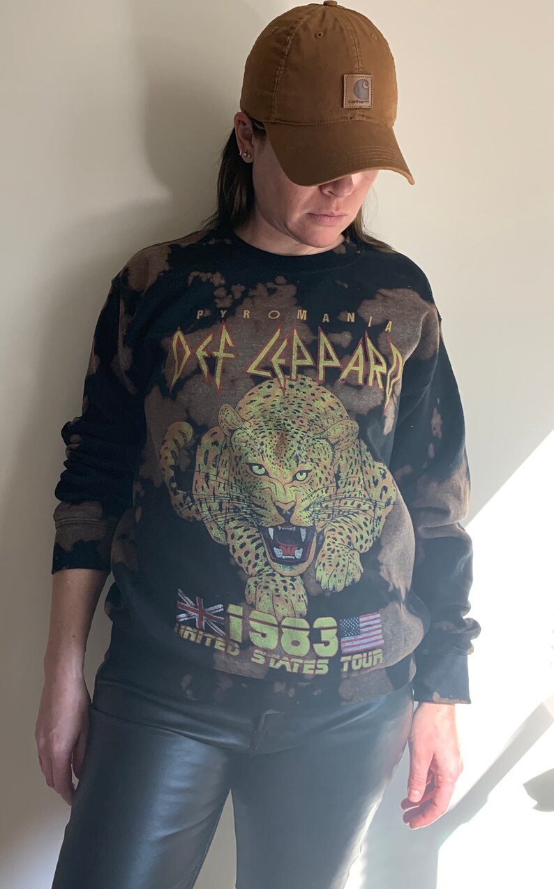 Puede incluir: Sudadera negra con efecto blanqueado que presenta un gr&aacute;fico de un leopardo con el texto "Pyromania Def Leppard 1983 United States Tour" impreso en la parte delantera. La sudadera la lleva puesta una persona que lleva una gorra de b&eacute;isbol marr&oacute;n.