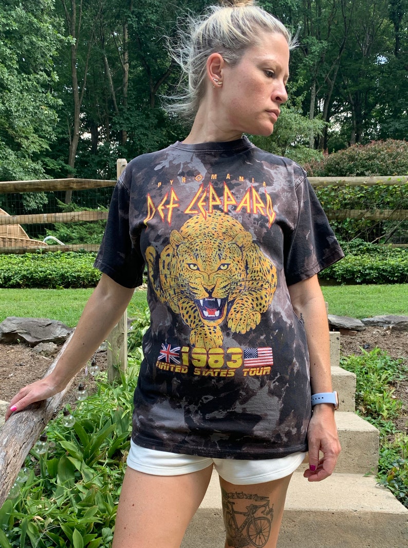 Puede incluir: Camiseta negra tie-dye con estampado de leopardo amarillo y naranja y el texto "Def Leppard Pyromania 1983 United States Tour" en amarillo y rojo.