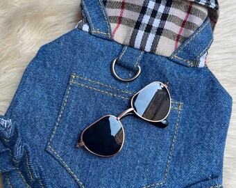 May include: Blue denim dog outfit with a plaid collar and a pocket. Gold-framed sunglasses with dark lenses rest on the outfit. The outfit has a ruffled hem and yellow stitching.
