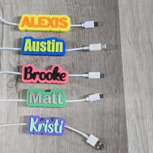 May include: A collection of personalized charging cable tags with names: Alexis, Austin, Brooke, Matt, and Kristi. Each tag is a different color, with the name in a contrasting color. The cables have USB-C and USB-A connectors.