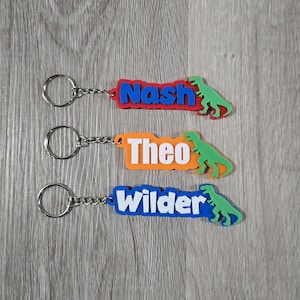 May include: Three personalized keychains with names "Nash", "Theo", and "Wilder". Each keychain features a rectangular tag with a different color and a green dinosaur silhouette. The names are in white, with a silver key ring.