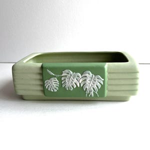 May include: A rectangular light green ceramic planter featuring horizontal ribbed detailing along the sides and a centered, darker green rectangular plaque adorned with a white, raised relief design of three monstera leaves on a vine.
