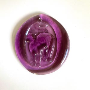 May include: A purple glass pendant with a raised elephant design. The pendant is teardrop-shaped with a hole at the top for hanging. The elephant is a darker shade of purple than the pendant's outer edge.