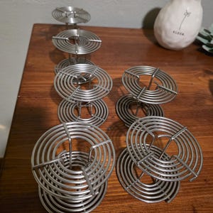 May include: A collection of silver-colored film developing reels. The reels are made of thin, circular metal wires, stacked in a spiral design. They are arranged on a dark wooden surface, with a white vase in the background.