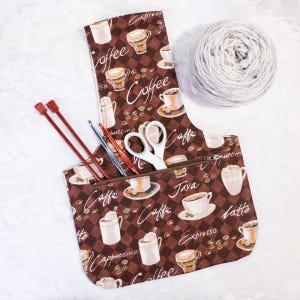 May include: A brown fabric project bag with a zippered pocket and a coffee cup pattern. The bag is filled with knitting needles, a crochet hook, and a pair of scissors. The text on the bag reads "Coffee", "Caffe", "Java", "Espresso", "Cappuccino", "Latte", and "Buccino".