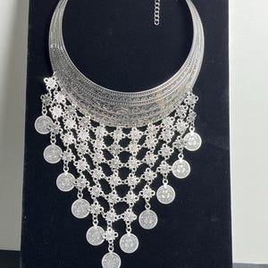 May include: A silver-toned statement necklace with a wide, crescent-shaped collar. The collar features intricate etched patterns and connects to a delicate chain. Below, a series of interconnected floral-shaped links support dangling coin-like pendants, creating a layered effect.