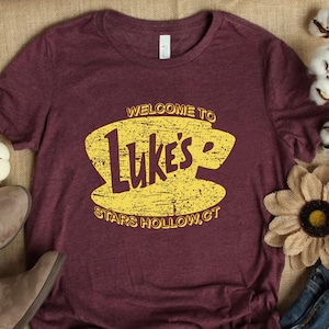 May include: A maroon t-shirt with a yellow graphic of a coffee cup and the text "Welcome to Luke's Stars Hollow, CT".