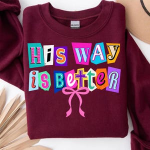 May include: A maroon sweatshirt with the text "His Way is Better" in a colorful, cut-out style. The text is in various colors, including pink, blue, orange, and yellow. A pink bow is at the bottom of the text.
