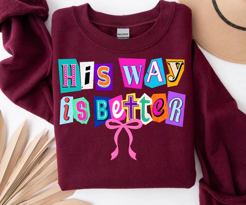 May include: A maroon sweatshirt with the text "His Way is Better" in a colorful, cut-out style. The text is in various colors, including pink, blue, orange, and yellow. A pink bow is at the bottom of the text.