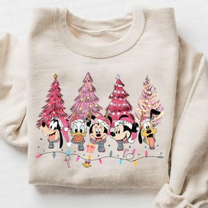 May include: A beige sweatshirt with a Christmas-themed design featuring Mickey Mouse, Minnie Mouse, Donald Duck, Goofy, and Pluto standing in front of pink and white Christmas trees. The characters are wearing Santa hats and holding gifts.